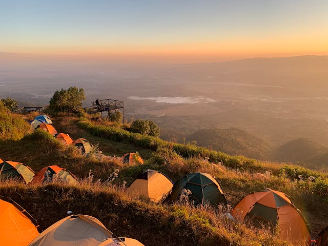 Zona de acampada con tiendas de campaña en una ladera de montaña al atardecer, con vistas panorámicas a un valle y laguna, ideal para senderismo y turismo rural.