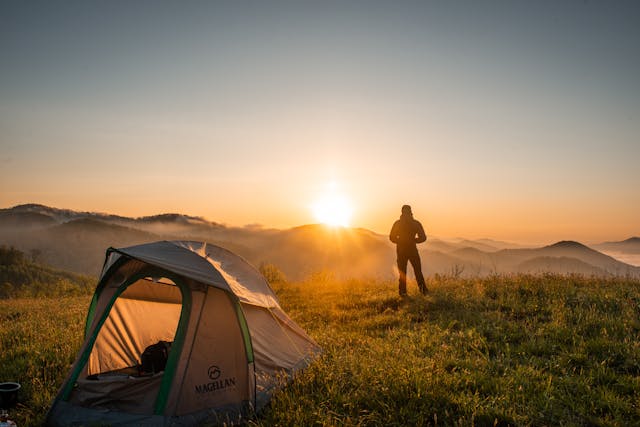 Persona de pie junto a tienda de campaña al amanecer en una pradera de montaña, con vistas panorámicas y luz dorada, ideal para camping y turismo de aventura.