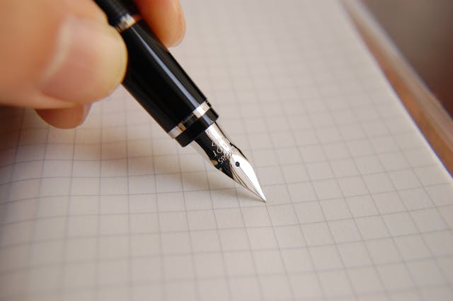 Mano escribiendo con pluma estilográfica sobre hoja de papel cuadriculado, ideal para representar redacción, escritura creativa o toma de notas.