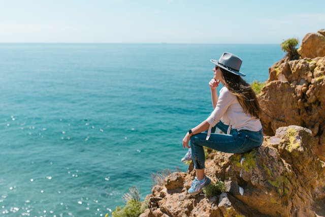 Mujer con sombrero sentada en un acantilado frente al mar en día soleado, contemplando el horizonte, ideal para ilustrar viajes, relax y turismo de costa.