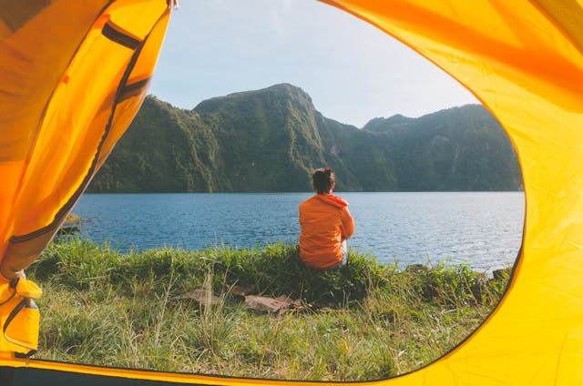Persona sentada frente a un lago de montaña vista desde el interior de una tienda de campaña amarilla, ideal para ilustrar experiencias de camping, desconexión y turismo de naturaleza.