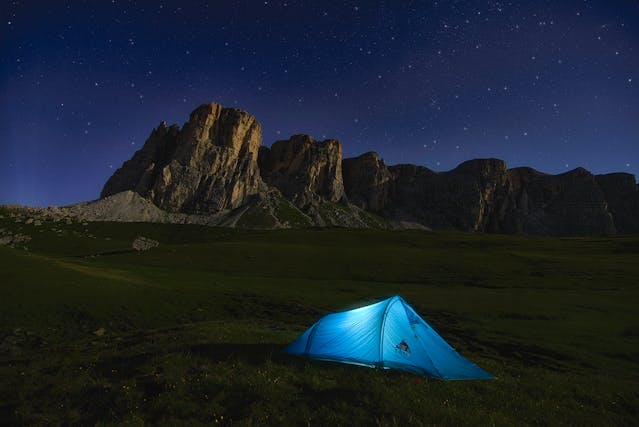 Tienda de campaña iluminada de noche bajo un cielo estrellado, en una pradera al pie de una cordillera rocosa, ideal para camping en alta montaña y astroturismo.
