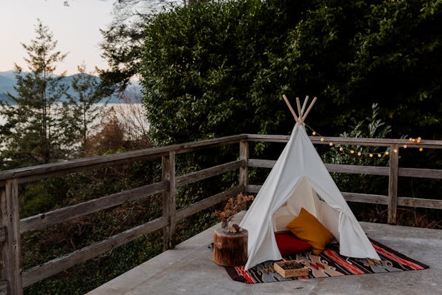 Pequeña tienda tipi decorativa sobre una terraza con vistas al bosque y lago, ideal para glamping, escapadas románticas o espacios de relax al aire libre.