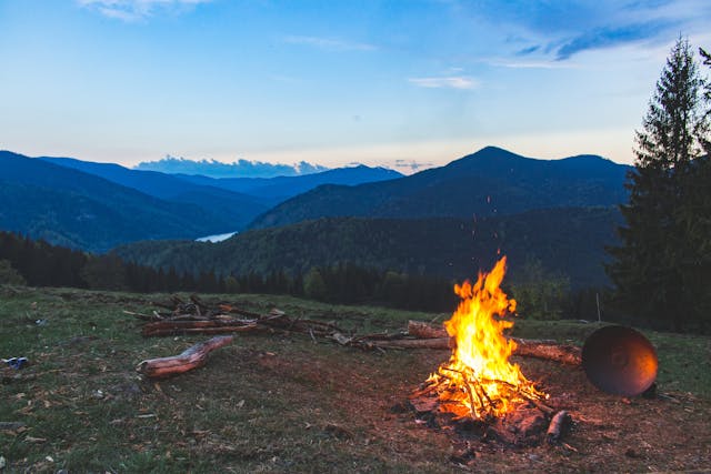 Fuego de campamento encendido al atardecer en una zona montañosa con vistas a valles y bosques, ideal para camping, naturaleza y experiencias al aire libre.